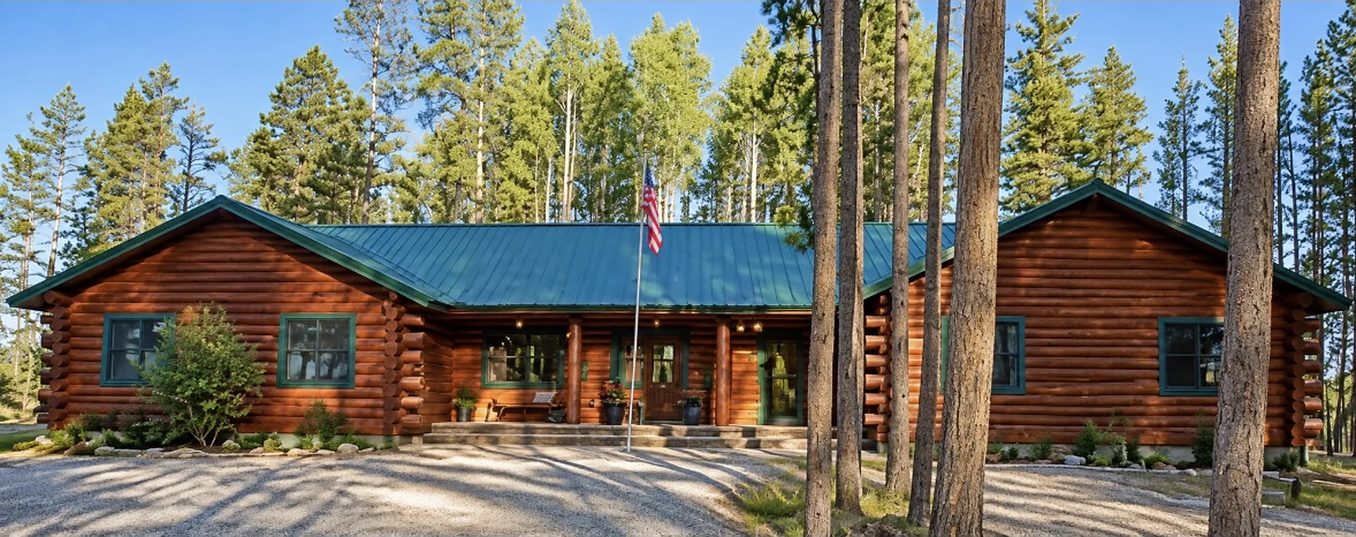Front of the log cabin with covered porch, teal metal roof and circle drive among lodgepole pines